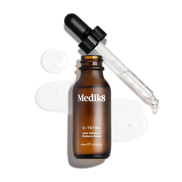 Een bovenaanzicht ("packshot") van een flesje Medik8 C-Tetra Lipid Vitamin C Radiance Serum van 30 ml. Het bruine glazen flesje is geopend en staat in het midden, met de zwarte pipetdop er diagonaal achter. Een helder, vloeibaar serum is artistiek rondom het flesje gespat op een strakke witte achtergrond, waarbij er ook enkele druppels zichtbaar zijn op de glazen pipet. De belichting is helder, wat zorgt voor zachte schaduwen aan de linkerkant en een klinische maar elegante uitstraling.-1