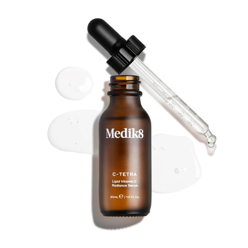 Een bovenaanzicht ("packshot") van een flesje Medik8 C-Tetra Lipid Vitamin C Radiance Serum van 30 ml. Het bruine glazen flesje is geopend en staat in het midden, met de zwarte pipetdop er diagonaal achter. Een helder, vloeibaar serum is artistiek rondom het flesje gespat op een strakke witte achtergrond, waarbij er ook enkele druppels zichtbaar zijn op de glazen pipet. De belichting is helder, wat zorgt voor zachte schaduwen aan de linkerkant en een klinische maar elegante uitstraling.