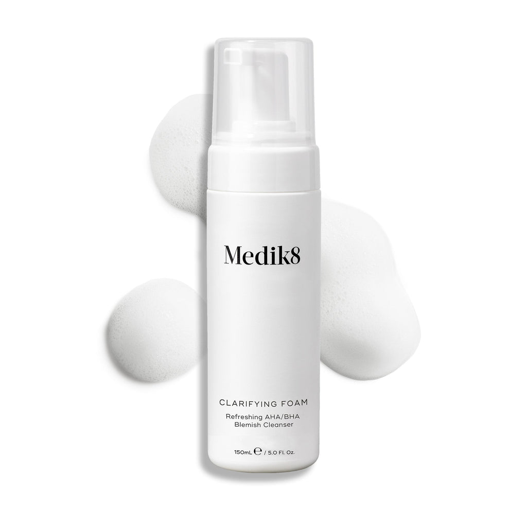 Een pompflacon Medik8 Clarifying Foam gepresenteerd op een strakke witte achtergrond met drie dotten wit, luchtig schuim. De tekst identificeert het product als een verfrissende AHA/BHA Blemish Cleanser met een inhoud van 150 ml.