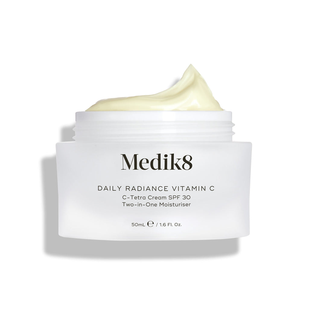 Een open glazen pot Medik8 Daily Radiance Vitamin C wordt getoond met een royale toef lichtgele crème bovenop. De tekst identificeert het product als een 50 ml C-Tetra Cream SPF 30, een twee-in-één moisturizer.