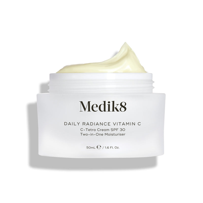 Een open glazen pot Medik8 Daily Radiance Vitamin C wordt getoond met een royale toef lichtgele crème bovenop. De tekst identificeert het product als een 50 ml C-Tetra Cream SPF 30, een twee-in-één moisturizer.