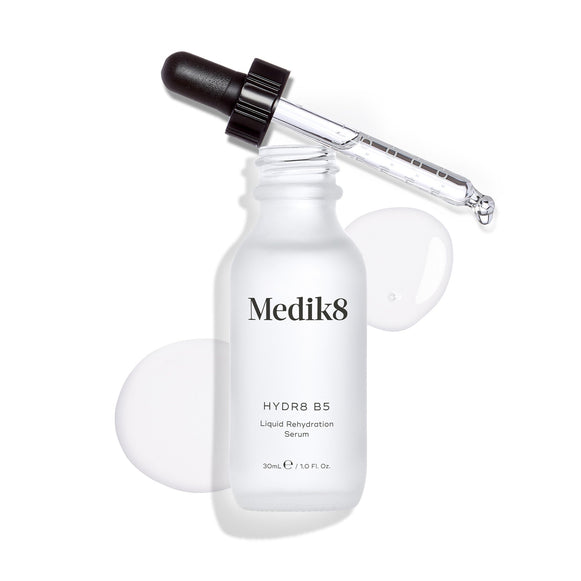 De fles Medik8 Hydr8 B5 serum staat tegen een strakke witte achtergrond met een heldere vloeistofplas en druppels, terwijl de pipet bovenop rust. De tekst identificeert het product als een 30 ml Liquid Rehydration Serum.-15