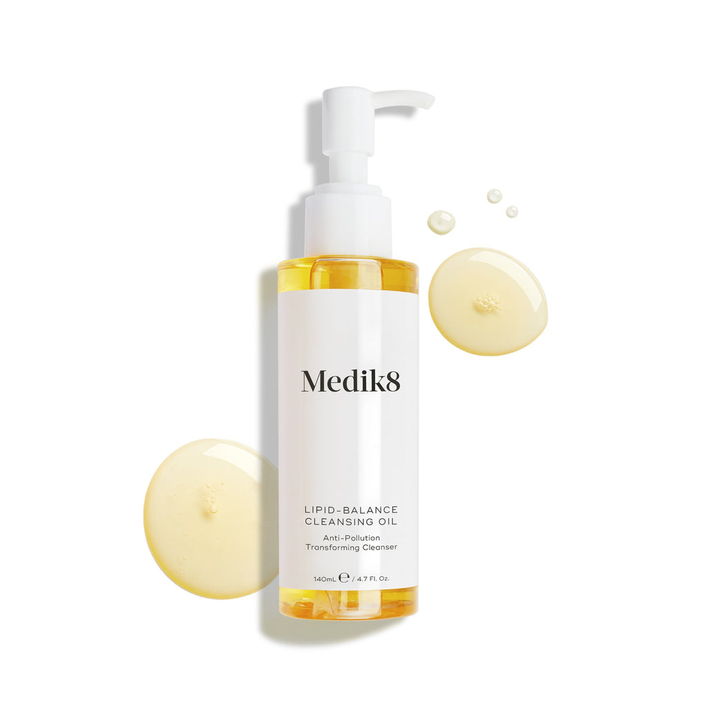 Een fles Medik8 Lipid-Balance Cleansing Oil staat tegen een witte achtergrond. Gouden oliedruppels omringen de fles en benadrukken de rijke, vloeibare textuur.