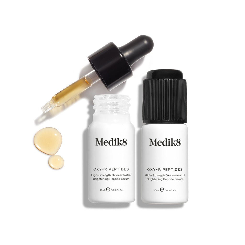 Een witte kartonnen verpakking van Medik8 Oxy-R Peptides naast twee kleine witte flesjes van het serum; één flesje met een zwarte pipet en de andere met een zwarte dop.