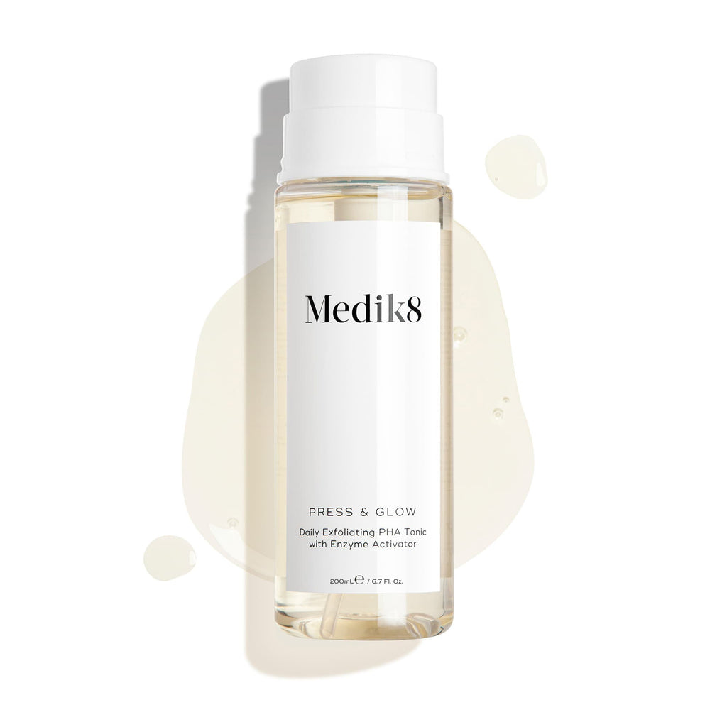 De Medik8 Press & Glow tonicfles staat tegen een strakke witte achtergrond met doorschijnende vloeistofdruppels en een plas, die de waterige textuur benadrukken. Tekst identificeert het product als een 200 ml Daily Exfoliating PHA Tonic met Enzyme Activator.