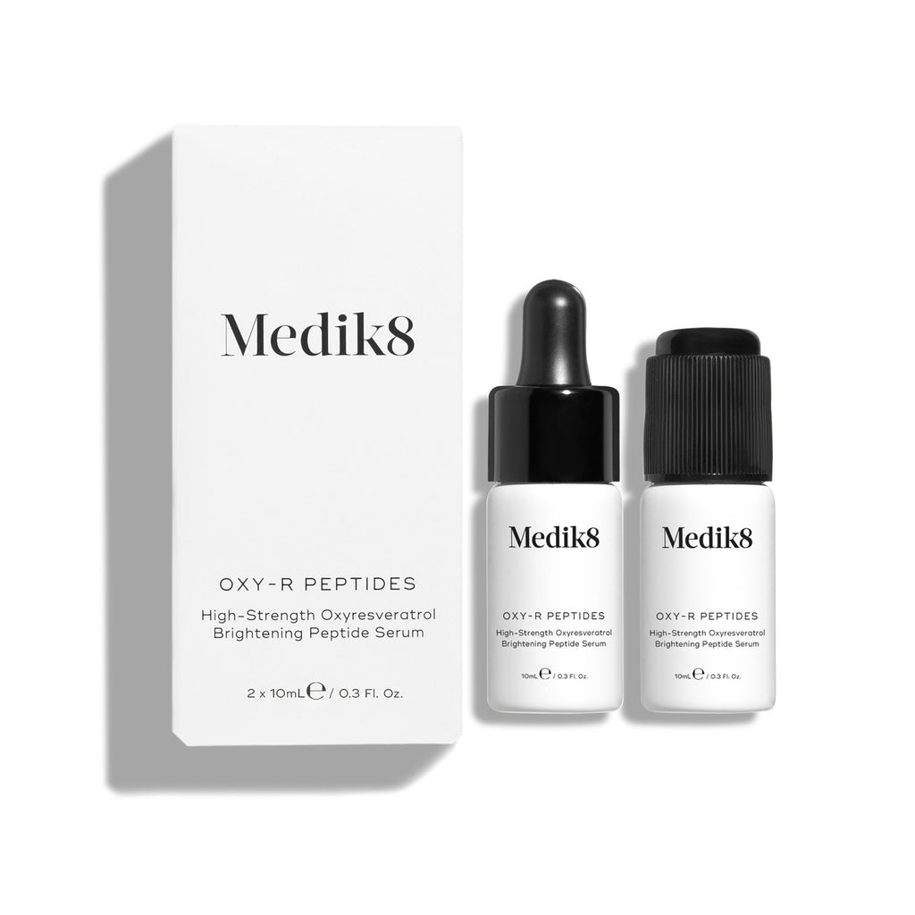 Twee kleine witte flesjes Oxy-R Peptides serum op een witte ondergrond. Ernaast ligt een zwarte pipet en zijn twee druppels geel serum zichtbaar op het oppervlak.