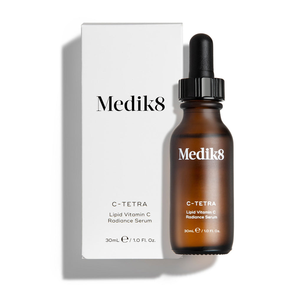 Een amberkleurig glazen flesje Medik8 C-Tetra lipid vitamin C radiance serum naast de bijbehorende witte kartonnen verpakking. Beide staan tegen een strakke witte achtergrond met een subtiele schaduw.