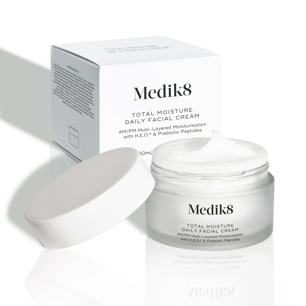 Een open, matglazen pot van Medik8 Total Moisture Daily Facial Cream met een witte deksel ernaast, waarbij de rijke witte crème zichtbaar is. De pot staat voor de witte kartonnen verpakking op een lichte achtergrond.