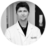 Een zwart-witportret van een Medik8-huidverzorgingswetenschapper en oprichter, gekleed in een labjas met merklogo, dat het onderzoek en de innovatie achter de formule vertegenwoordigt.